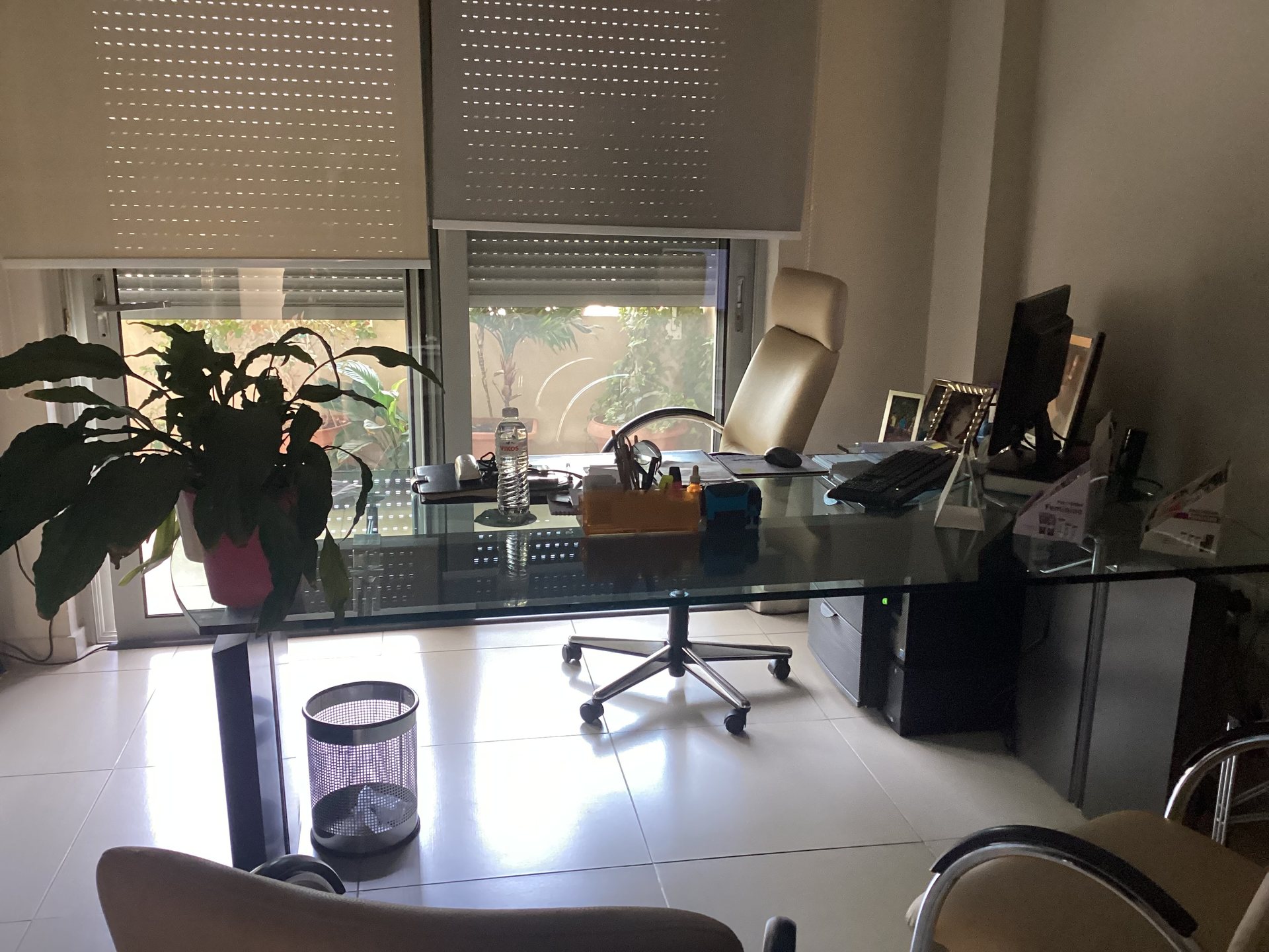 Γραφείο ιατρού με γυάλινο τραπέζι, υπολογιστή και φυτό μπροστά από τη μπαλκονόπορτα